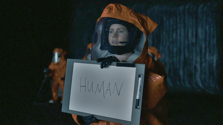 Louise wearin a hazmat suit and holding up a clipboard with the word human written on it