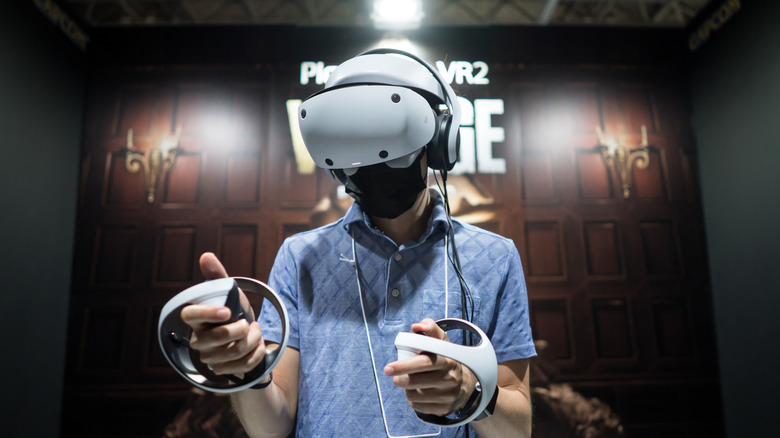 An individual in a blue shirt is utilizing a PlayStation 5 VR2 headset, the user is wearing the headset as they hold two white controllers in each hand