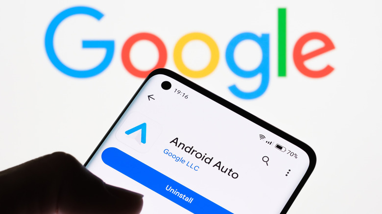 A smartphone being held in a user's left hand displays the Android App app page as the smartphone is being held against a white background displaying the Google logo