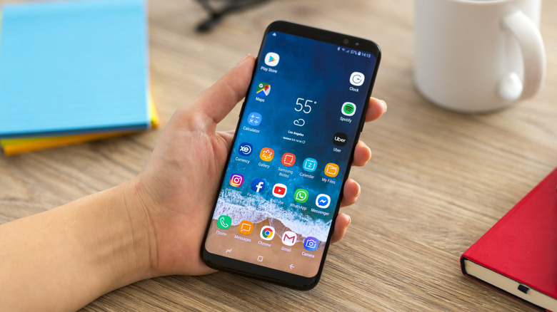 A Samsung Galaxy S9 displaying its Home screen as it sits in a user's left hand, the hand rests on a desk that has a stack of post-it notes, a coffee cup, and a notebook