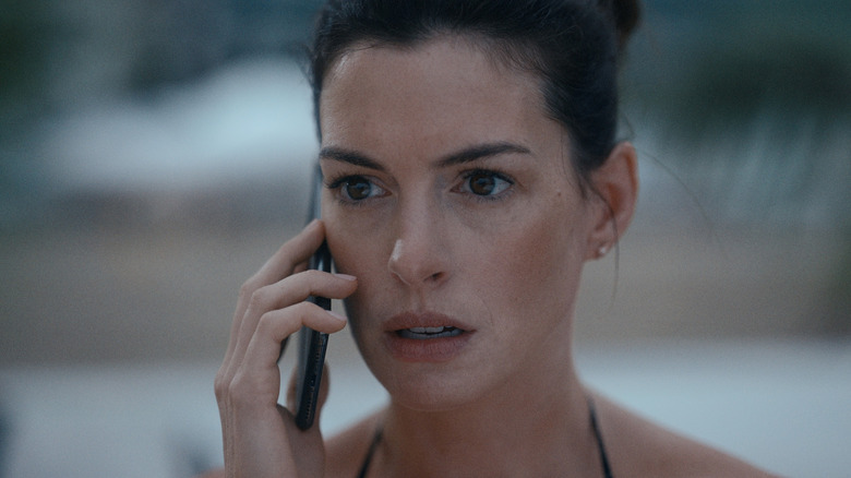 Rebekah (Anne Hathaway) looks shocked as she talks on the phone on the sand, her dark hair in a messy bun