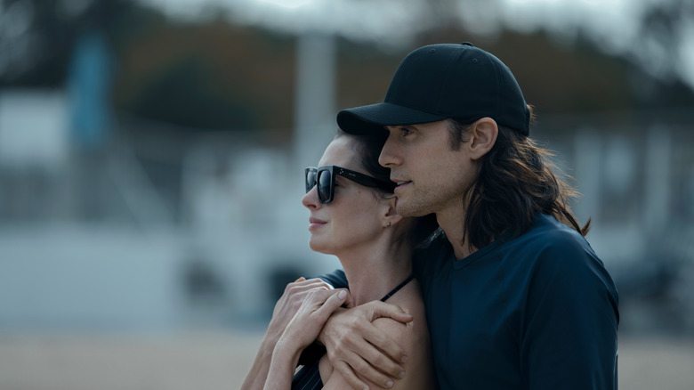 Adam Neumann (Jared Leto), clothed in dark shirt and black hat, clutches his wife Rebekah (Anne Hathaway) to him on the beach