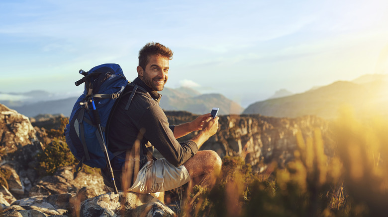 Young male hiker sitting down and smiling at camera with phone in hands