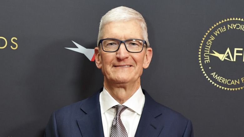 Tim Cook smiling, standing against a gray background.