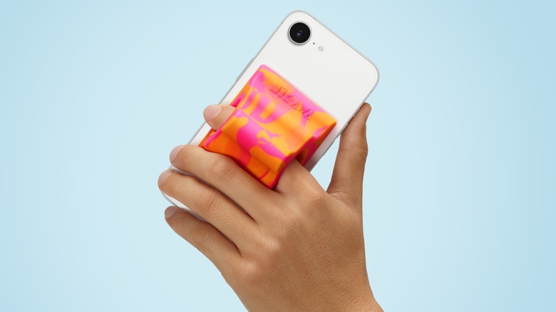 A hand holding a white iPhone that has the orange Hikawa Phone Grip & Stand attached to it