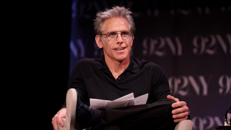 Ben Stiller, wearing aviator-style glasses, is seated on a stage with his legs crossed, several printed pieces of paper are in his lap.