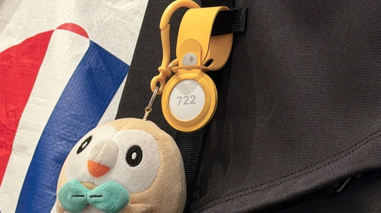 An AirTag attached to a backpack with the engraved numbers 722, alongside a plushie of the Pokémon Rowlet