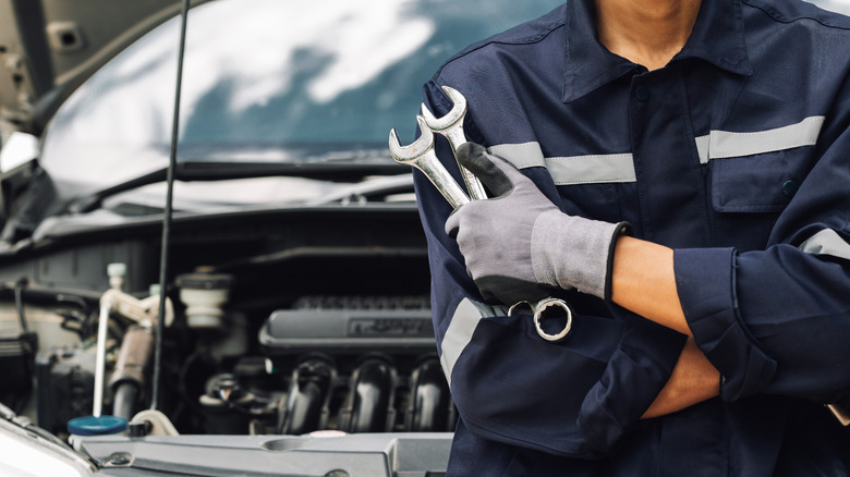 A car mechanic holding several tools. He stands in front of a car with its hood open.