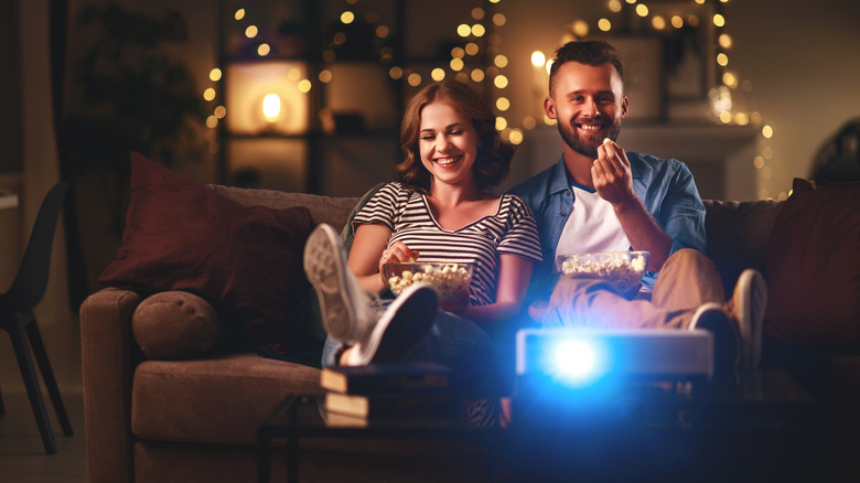 a couple eating popcorn on the couch with their home projector in front of them