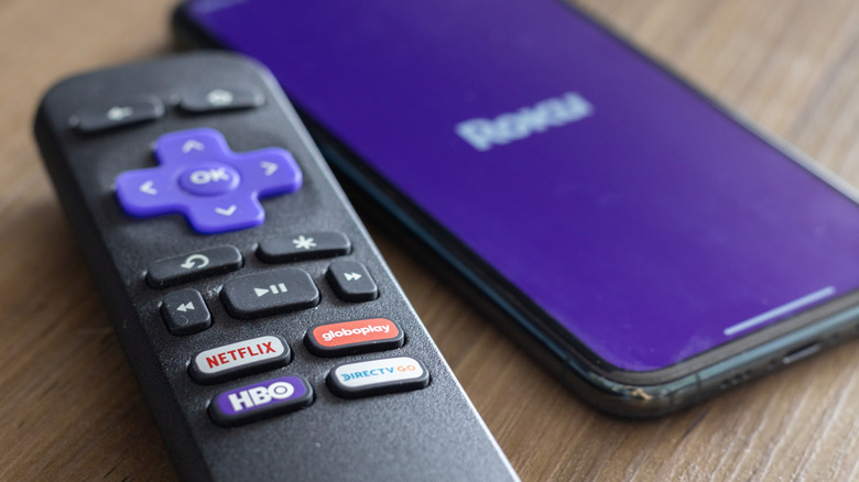 A Roku streaming device remote placed next to a phone with the Roku logo on its screen.