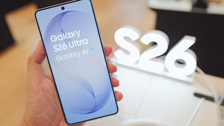 Hand holding a Samsung Galaxy S26 Ultra with the Galaxy AI screen shown beside an illuminated S26 sign.