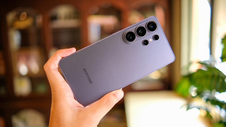 Hand holding a Samsung smartphone with the rear cameras facing forward in a warmly lit indoor setting.