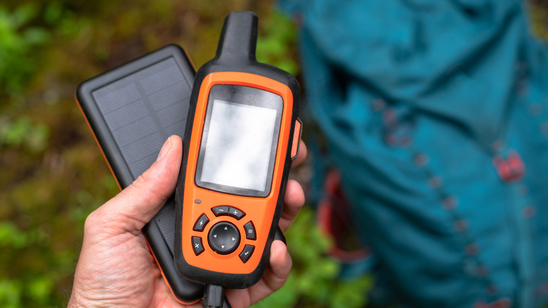 A hand holding a device on a solar-powered portable battery pack with camping gear in the background.