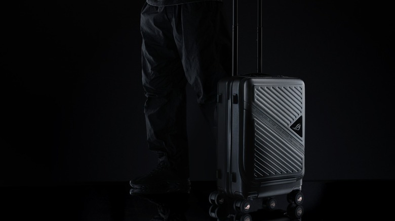 An individual in black holdings the handle of a ASUS ROG Archer Suitcase 20, both the individual and the suitcase are against a black background, and the image is cropped at the user's torso
