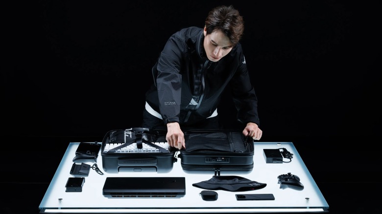 An individual in all black is packing a ASUS ROG Archer Suitcase 20, the suitcase is sitting on an illuminated table surrounded by several accessories, and the theme of the image is black