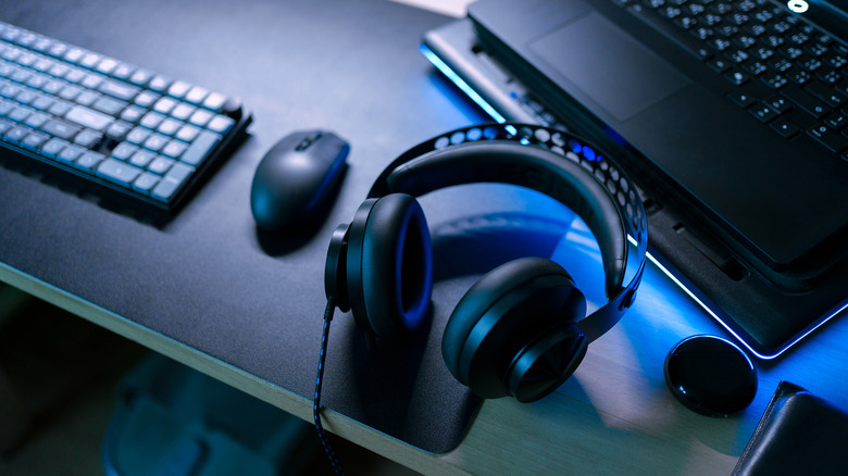 A pair of black headphones sitting on a desk next to a mouse, keyboard, and gaming laptop