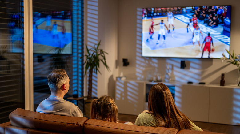 A family watching a basketball game on a wall-mounted living room TV.