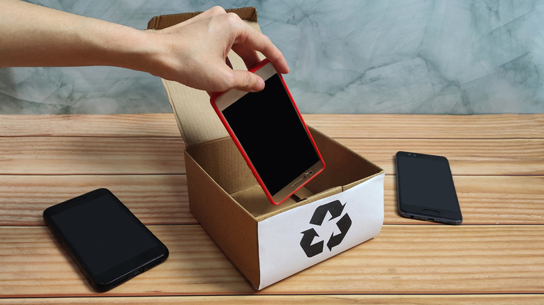 person placing an old phone in a recycle box with two other phones kept on the side on a wooden table