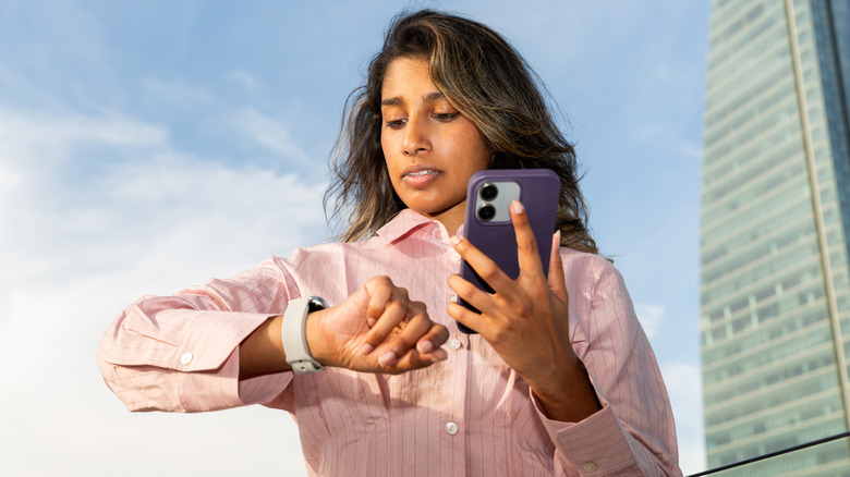 A young woman outside looking at her smartwatch, her phone in the other hand.