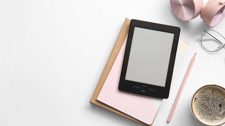 An e-reader placed on a set of books next to a cup of coffee, pencil, and pair of headphones