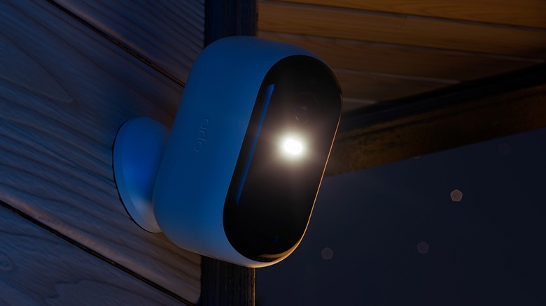 An Arlo Essential Security Camera 2K mounted to a wooden exterior of a house with its spotlight turned on