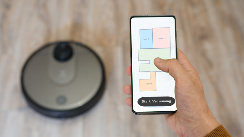 A robot vacuum on the floor with a hand holding a phone running its accompanying app.