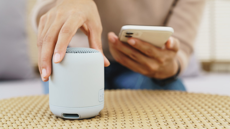 A person holding their phone in one hand, using the other to place a Bluetooth speaker on a table