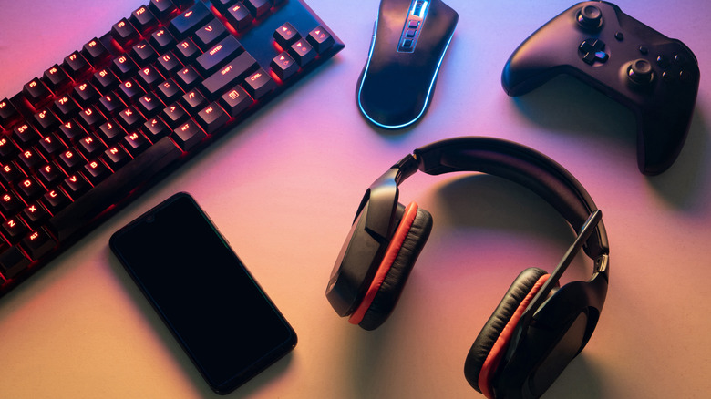 Various gaming gear sitting on a table including a keyboard, controller, mouse, phone, and headset