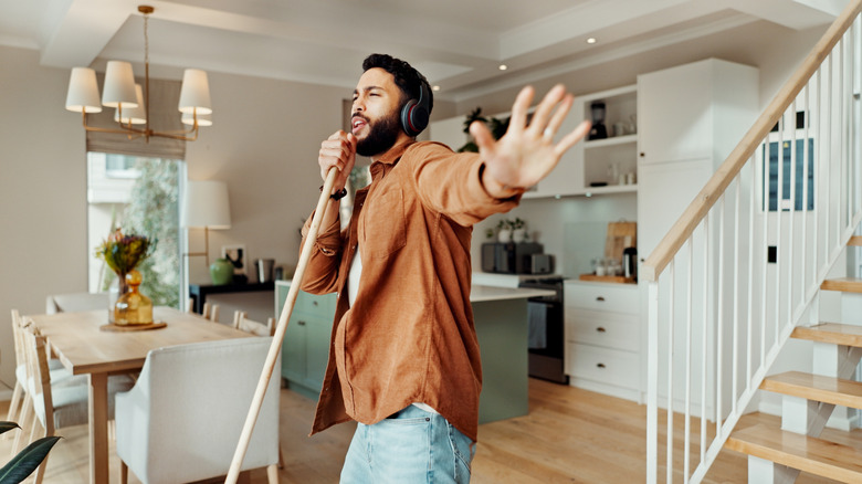 A man stands in a kitchen, and uses a mop handle as a faux microphone.