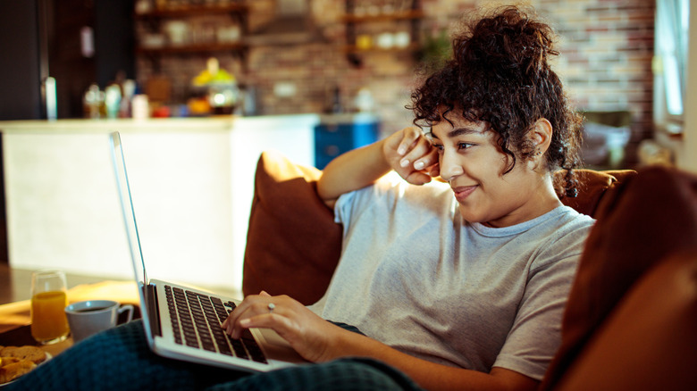 A young woman lounging on a sofa smiling at her laptop with her hand on the keyboard.