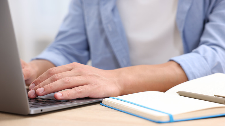 A person using a laptop with a notebook next to them.