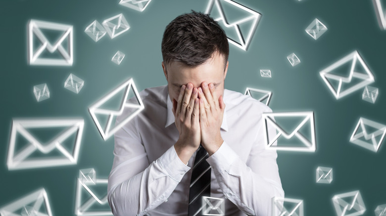 A man in a dress shirt and tie buries his face in his hands as he superimposed email icons swirl all around him