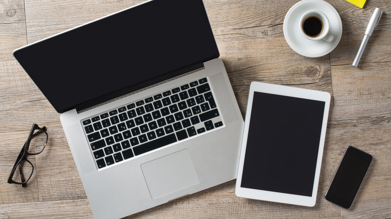 Laptop, tablet, and smartphone placed on a wooden table alongside glasses, pen and a cup of coffee