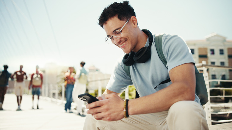 A young person texting on a smartphone outdoors with people in the background.