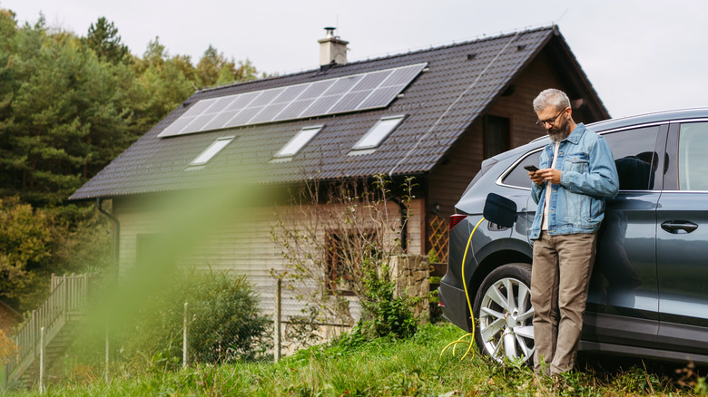 A man looking at his phone while charging an electric car in front of a building with solar panels on the roof.