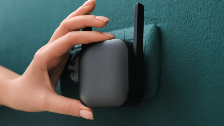 A person with well-manicured nails plugging a black Wi-Fi repeater into an open wall socket.