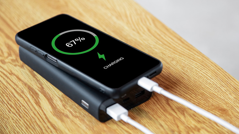 A black smartphone charging atop a black portable battery pack on a beige wooden surface.
