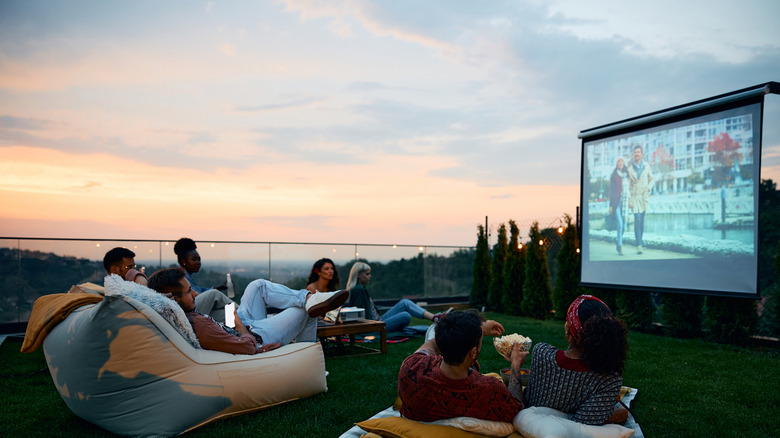 group of friends sitting outdoors watching a movie on a projector screen as the sun sets in the background