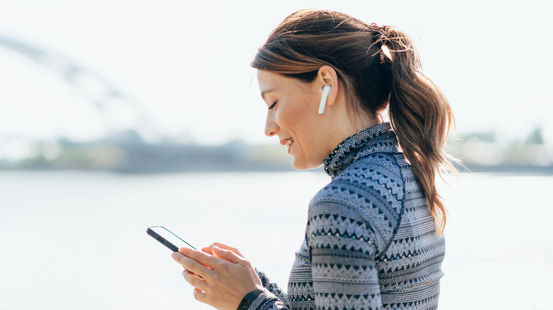 A woman standing in front of a river, wearing wireless earbuds.