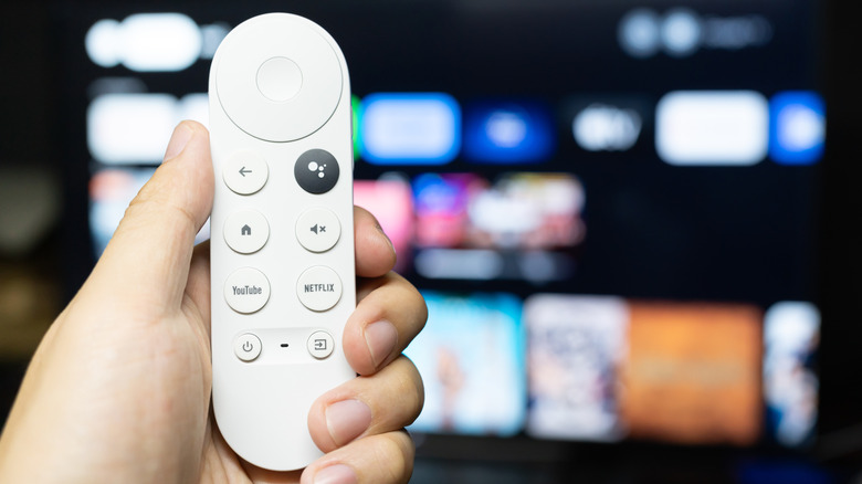 An individual holds a Google TV remote in their left hand as it's held in front of a heavily blurred TV that seems to be displaying a smart TV home screen