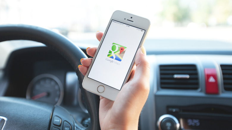 A closeup of a hand in front of the steering steering wheel in a vehicle, the hand is holding a white iPhone displaying the Google Maps icon