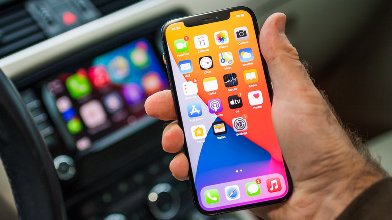 A closeup of a hand holding an iPhone 12 with CarPlay active on a vehicle's infotainment system in the background