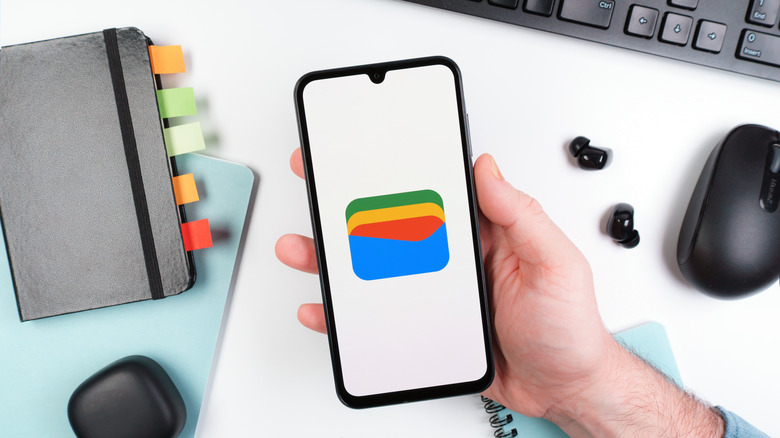 A person holding a smartphone with the Google Wallet app open. Various desk items are in the background, including a notebook, earbuds, keyboard, and mouse.