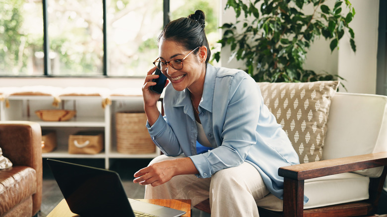 A woman smiling while talking on a smartphone and looking at a laptop