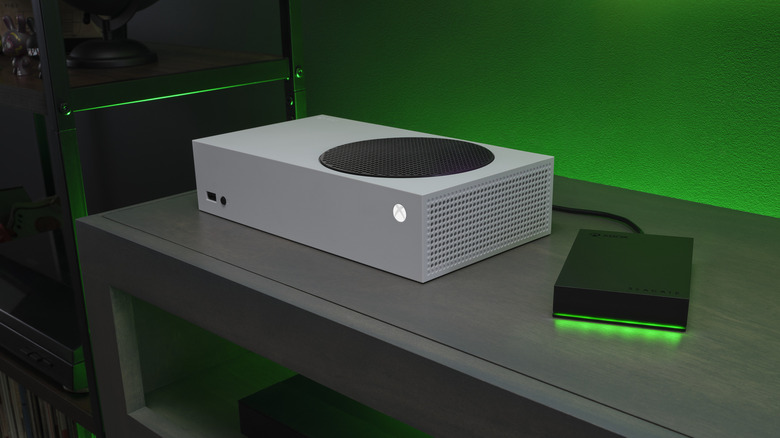 A white Xbox Series X on a gray surface with a black game drive attached with a wire