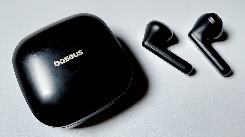 A pair of Baseus earbuds, next to their charging case, on a pale background.