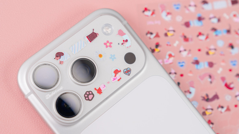 Apple iPhone 17 Pro camera module with small decorative stickers on a pink surface, with a blurred sticker sheet in the background creating a playful customization concept