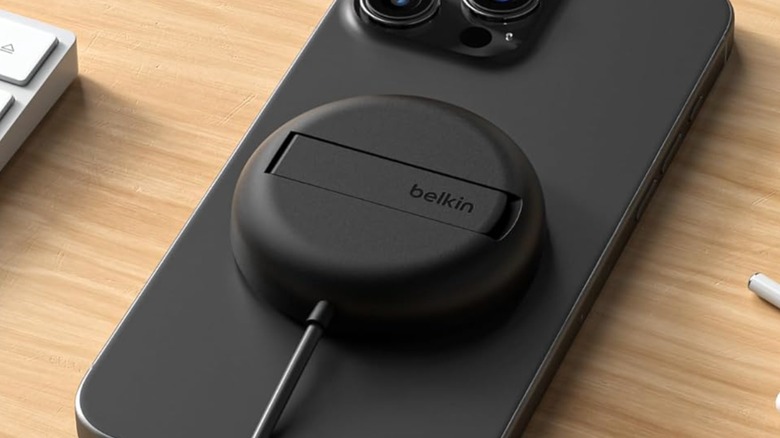 A closeup of the Belkin Magnetic Wireless Charging Pad with Kickstand as it sits atop a black iPhone, both items are sitting on a desk between a AirPods and a keyboard