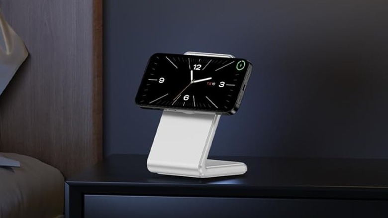 A white Enyneo MagSafe iPhone Charger Stand sitting on a black table in what appears to be a bedroom, the attached iPhone is displaying a clock