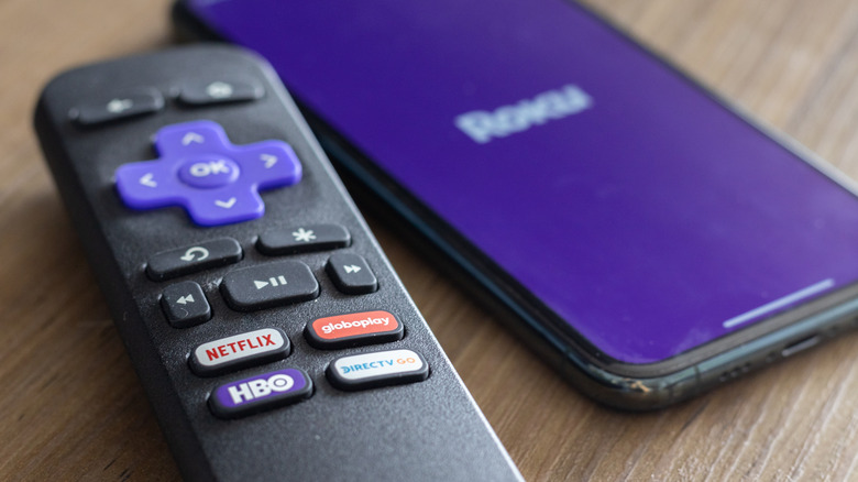 Roku remote up close with app buttons and the Roku logo on a smartphone in background.